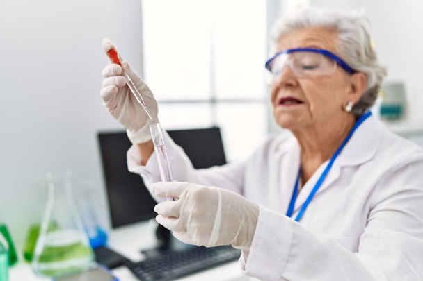 Ηλικιωμένη γκριζομάλλα γυναίκα που φοράει επιστημονική στολή χρησιμοποιώντας πιπέτα και δοκιμαστικό σωλήνα στο εργαστήριο - Φωτογραφία, εικόνα