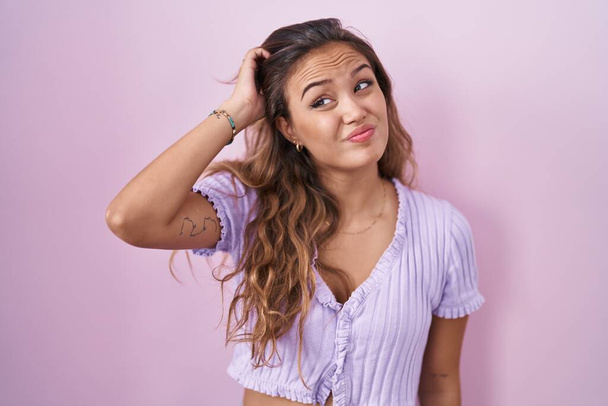 Young hispanic woman standing over pink background confuse and wondering about question. uncertain with doubt, thinking with hand on head. pensive concept.  - Φωτογραφία, εικόνα