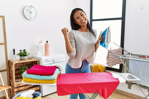 Νεαρή Ισπανίδα σιδερώνει ρούχα στο σπίτι δείχνοντας τον αντίχειρα στο πλάι χαμογελώντας χαρούμενη με ανοιχτό στόμα.  - Φωτογραφία, εικόνα