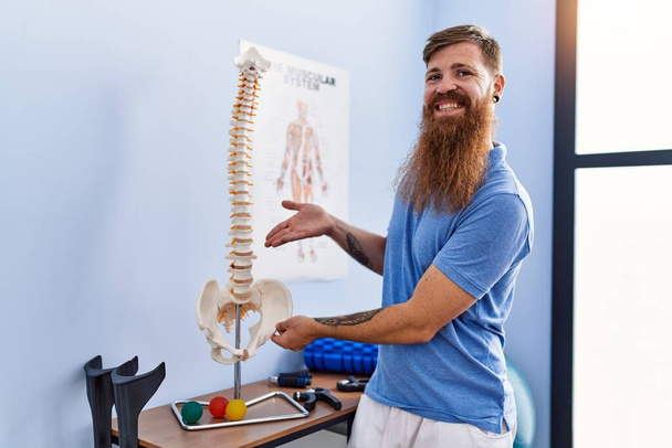 Joven pelirrojo vistiendo uniforme fisioterapeuta apuntando al modelo anatómico de columna vertebral en clínica de fisioterapia - Foto, Imagen