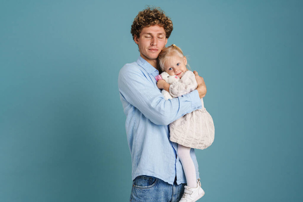 Curly young man holding and hugging his daughter on camera isolated over blue background - Foto, Imagem