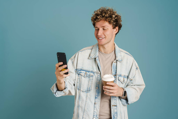 Young curly man using mobile phone while drinking coffee isolated over blue background - Foto, Imagem
