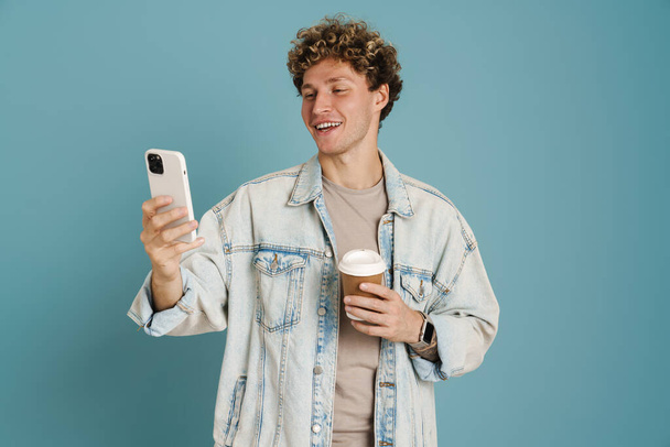Young curly man using mobile phone while drinking coffee isolated over blue background - Photo, Image