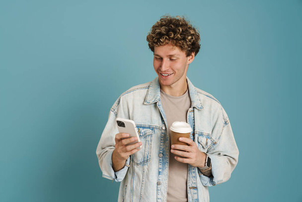 Young curly man using mobile phone while drinking coffee isolated over blue background - Φωτογραφία, εικόνα