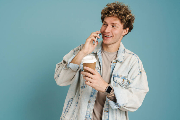 Young curly man drinking coffee while talking on mobile phone isolated over blue background - Fotó, kép