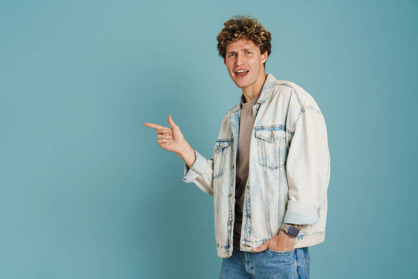 Young curly man wearing jacket smiling and pointing finger aside isolated over blue background - Photo, Image