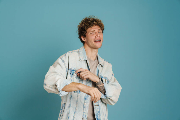 Young curly man wearing jacket singing and dancing isolated over blue background - Foto, Imagen