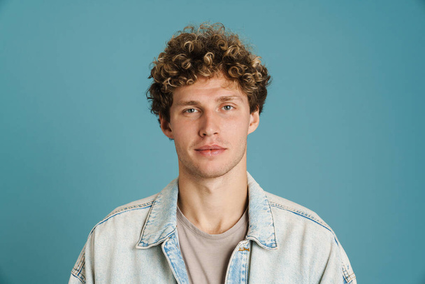 Young curly man wearing jacket posing and looking at camera isolated over blue background - Foto, afbeelding