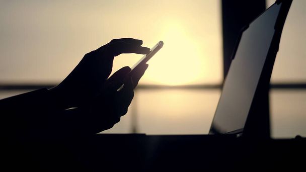 dark silhouette of female hands, typing something in mobile, next is laptop on table. against background of a large office window, at sunset, in the rays of light. close up. High quality photo - Φωτογραφία, εικόνα