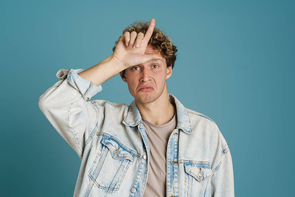 Young curly man grimacing and pointing finger upward isolated over blue background - 写真・画像