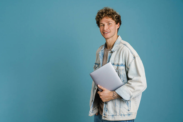 Young man wearing denim jacket smiling while posing with laptop isolated over blue background - Fotoğraf, Görsel