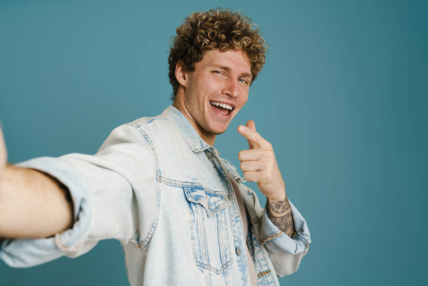 Young excited man pointing finger away while taking selfie photo isolated over blue background - Fotoğraf, Görsel