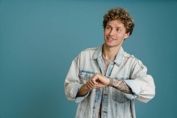 Young blonde man wearing denim jacket using wristwatch and smiling isolated over blue background - Foto, imagen