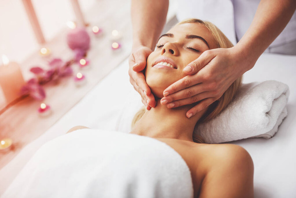 Beautiful blonde girl is lying with closed eyes at spa while a massage therapist is massaging her head - Fotografie, Obrázek