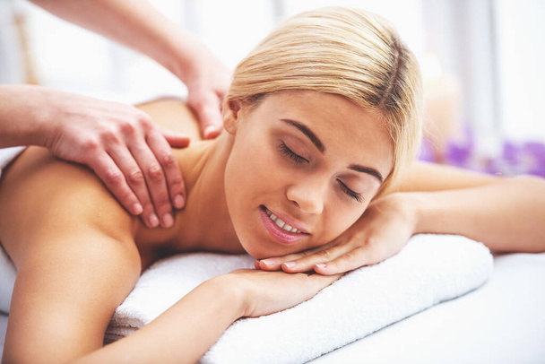 Beautiful blonde girl is smiling, lying with closed eyes on front of her body while a massage therapist is massaging her shoulders - Photo, Image