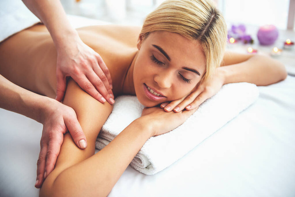 Mooi blond meisje lacht, liggen met gesloten ogen op de voorkant van haar lichaam, terwijl een massagetherapeut is haar rug masseren - Foto, afbeelding