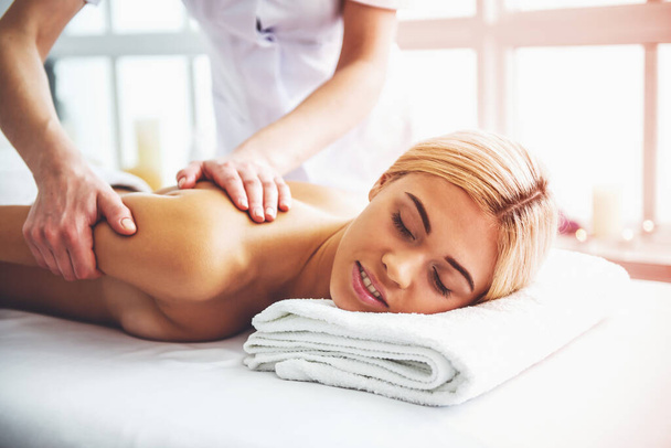 Beautiful blonde girl is smiling, lying with closed eyes on front of her body while a massage therapist is massaging her back - Fotografie, Obrázek