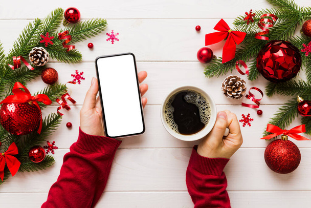 Powyżej widok kobiecej ręki trzymającej smartfon z gorącą filiżanką kawy. Dekoracja świąteczna na kolorowym tle. - Zdjęcie, obraz
