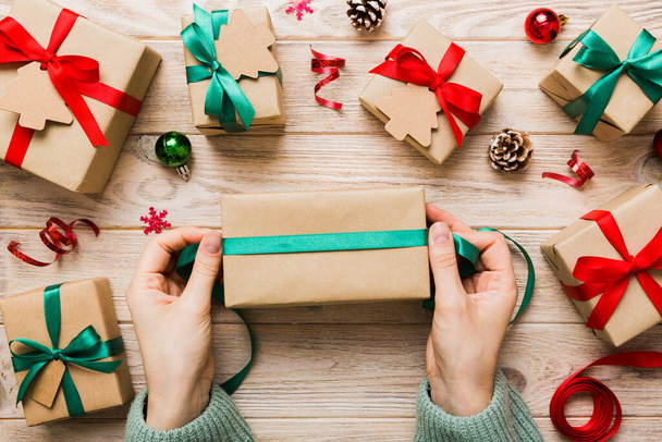 Γυναίκα συσκευάζει δώρο έκπληξη για τις διακοπές των Χριστουγέννων, κάθεται στο χώρο εργασίας με εργαλεία χειροτεχνίας, κορυφαία άποψη. Γυναίκα περιτύλιγμα χριστουγεννιάτικο δώρο στο χρωματιστό τραπέζι. - Φωτογραφία, εικόνα