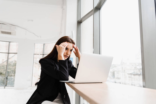 Woman manager with strong headache working on laptop online remotely in white office. Sad girl with laptop got migraine touches her head because of pain - Photo, Image