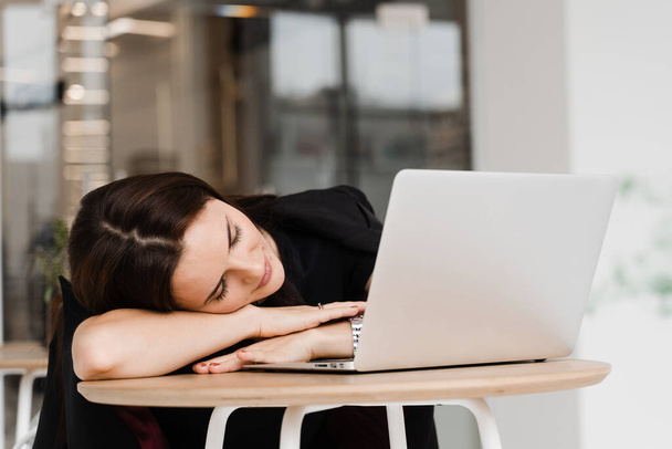 Девушка с закрытыми глазами отдыхает на работе и спит и расслабляется за ноутбуком на рабочем месте из-за сверхурочной работы - Фото, изображение