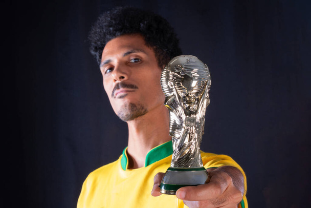 Jugador brasileño de fútbol negro sosteniendo trofeo ganador y celebrando, aislado sobre fondo negro. - Foto, Imagen