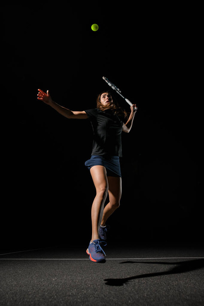 view on female tennis player with tennis racket in her hand bouncing to hit yellow tennis ball. Dark background. Movement, sport, healthy lifestyle concept. - Photo, image