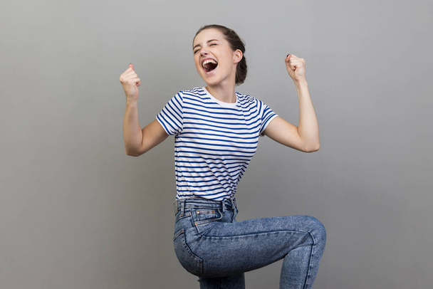 I am champion. Portrait of woman wearing striped T-shirt screaming for joy, raising high her fists, celebrating successful winning, incredible victory. Indoor studio shot isolated on gray background. - 写真・画像