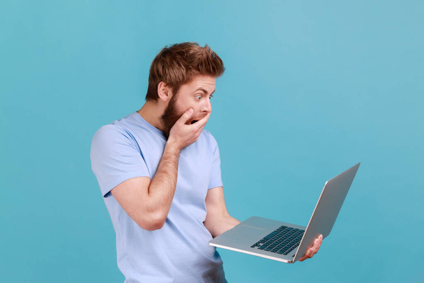 Portrait of handsome astonished surprised bearded man looking at laptop display with big eyes shocked with content, covered mouth with palm. Indoor studio shot isolated on blue background. - Φωτογραφία, εικόνα