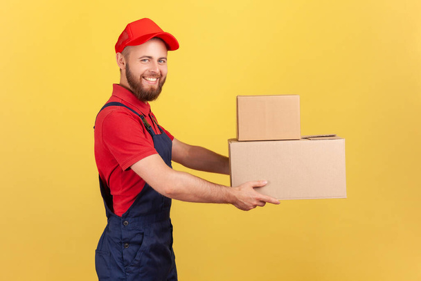 Side view of happy deliveryman holding two cardboard boxes, delivering order door-to-door, shipment and cargo transportation service. Indoor studio shot isolated on yellow background. - Zdjęcie, obraz