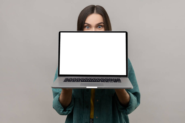 Positive unknown woman hiding half of face behind laptop with white empty display, looking at camera with big surprised eyes, wearing casual jacket. Indoor studio shot isolated on gray background. - Photo, image
