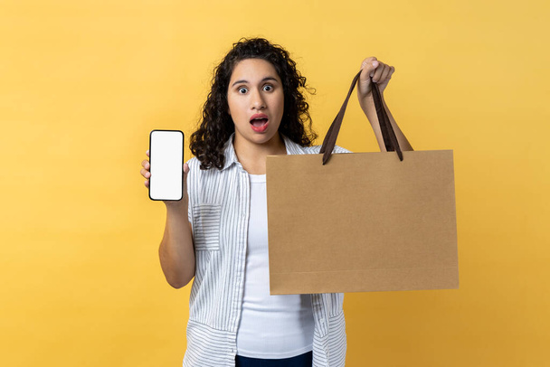 Portrait of surprised astonished woman with dark wavy hair holding paper shopping bag and mobile phone with empty screen, copy space for promotion. Indoor studio shot isolated on yellow background. - Фото, изображение
