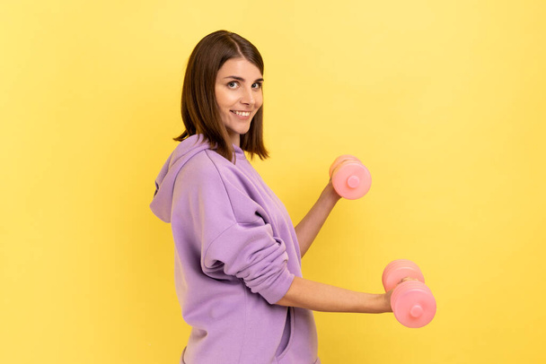 Side view of woman standing doing fitness exercises with pink dumbbells, training hands, looking at camera with smile, wearing purple hoodie. Indoor studio shot isolated on yellow background. - Zdjęcie, obraz
