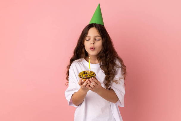 Cute charming little girl in green party cone on head holding cake and blowing candle, making wish, expressing happiness, keeps eyes closed. Indoor studio shot isolated on pink background. - Foto, imagen