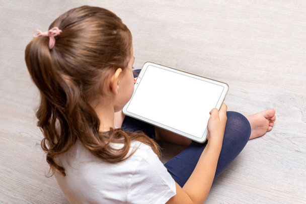 Bambina seduta sul pavimento e con in mano un tablet bianco con schermo isolato per promuovere videogiochi, siti web o app - Foto, immagini