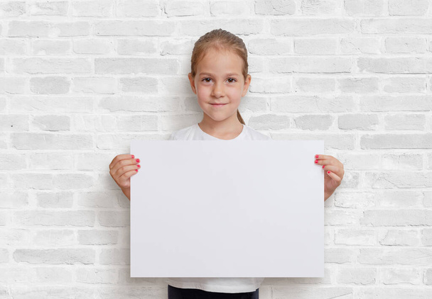 Girl holding a poster in a horizontal position. Blank paper for design presentation. White brick wall in background - Zdjęcie, obraz