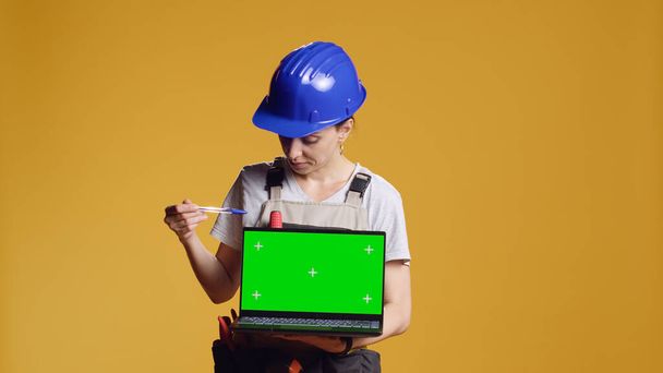 Portrait d'une bricoleuse tenant un ordinateur portable avec écran vert sur fond jaune, à l'aide d'un gabarit de clé chromatique vierge avec un espace de copie de maquette isolé. Rénovation avec ordinateur. - Photo, image