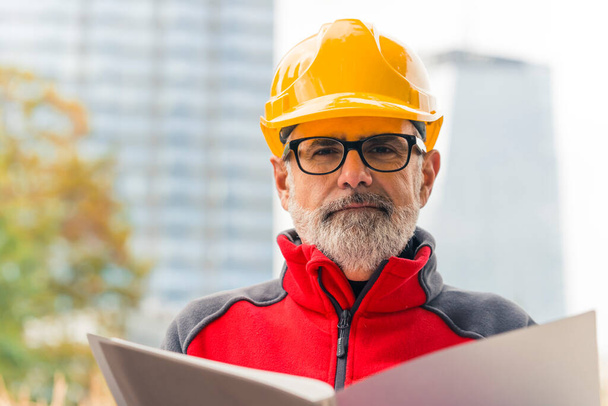 Middle-aged male contractor with beard wearing glasses and hard hat holding folder of documents looking into camera. Man in manager position. Horizontal outdoor shot . High quality photo - Zdjęcie, obraz