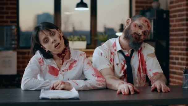 Bezmyślne zombie z biura jedzące mózgi, siedzące przy biurku, machające rękami przed kamerą. Przerażające potwory z głębokimi i krwawymi bliznami i ranami siedzące w firmowym miejscu pracy podczas warczenia - Materiał filmowy, wideo
