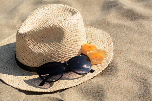 Hat with beautiful sunglasses and flowers on sand, closeup - Фото, изображение