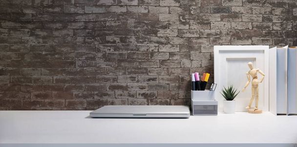Laptop komputer, ramka obraz, książki i uchwyt na ołówek na białym stole z cegły ściany. - Zdjęcie, obraz
