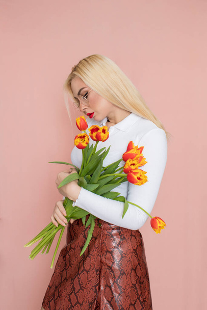 People emotions concept. Pretty adult woman stands indoor wears white shirt and brown snake skirt, round glasses isolated on pink background. Holding tulips in her hands. Springtime - Zdjęcie, obraz