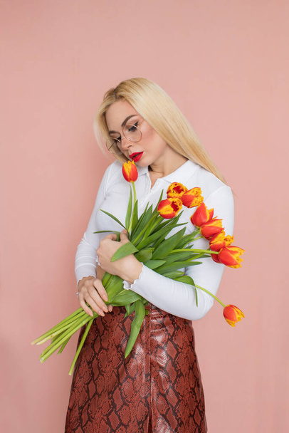 People emotions concept. Pretty adult woman stands indoor wears white shirt and brown snake skirt, round glasses isolated on pink background. Holding tulips in her hands. Springtime - 写真・画像