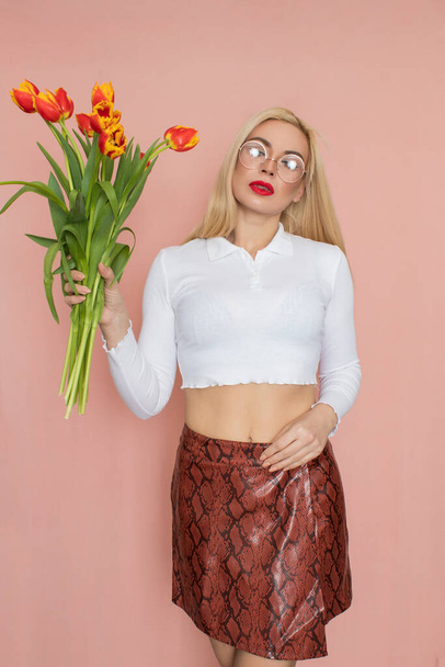 People emotions concept. Pretty adult woman stands indoor wears white shirt and brown snake skirt, round glasses isolated on pink background. Holding tulips in her hands. Springtime - Valokuva, kuva