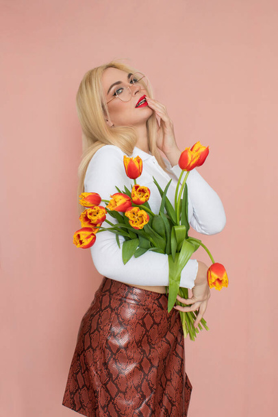 People emotions concept. Pretty adult woman stands indoor wears white shirt and brown snake skirt, round glasses isolated on pink background. Holding tulips in her hands. Springtime - Фото, зображення