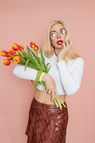 People emotions concept. Pretty adult woman stands indoor wears white shirt and brown snake skirt, round glasses isolated on pink background. Holding tulips in her hands. Springtime - Photo, Image