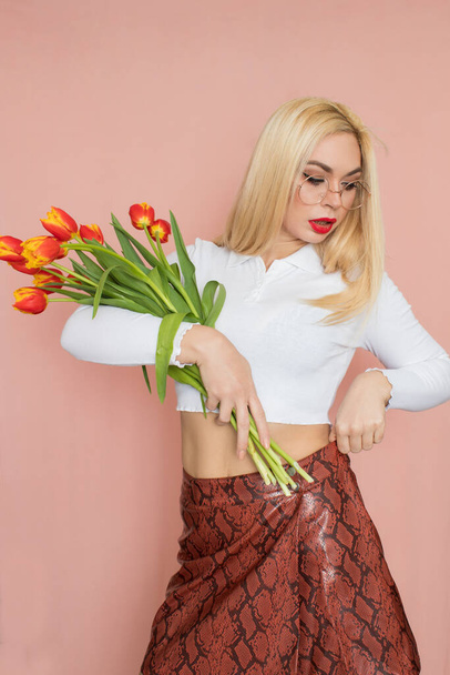 People emotions concept. Pretty adult woman stands indoor wears white shirt and brown snake skirt, round glasses isolated on pink background. Holding tulips in her hands. Springtime - Foto, Bild