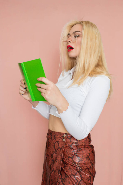 People emotions concept. Pretty adult woman stands indoor wears white shirt and brown snake skirt, round glasses isolated on pink background. Thinking about new ideas with book and pencil in her hands - Photo, image