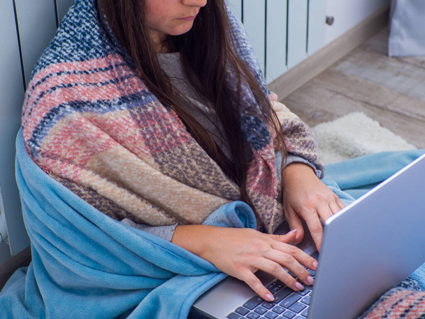 Žena sedí u baterie doma a pracuje s laptopem. Teplě oblečená žena je rozrušená z vysoké ceny topení. Drahé energetické zdroje, energetická krize v Evropě. Online vzdělávání, práce - Fotografie, Obrázek