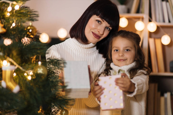 Loving happy young caucasian mother at fir tree with small cute daughter holding Christmas gifts, presents. Smiling mom and girl child celebrate New Year holidays at home. - Photo, image